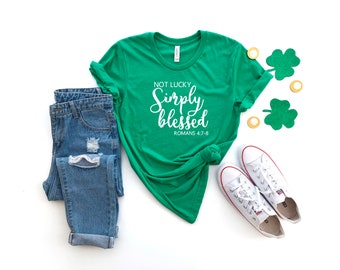 May include: Green t-shirt with white text that reads "Not Lucky Simply Blessed Romans 4:7-8". The shirt is paired with blue jeans and white sneakers. There are green shamrocks and gold coins on the white background.