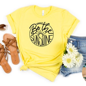 May include: Yellow t-shirt with the text "Be the Sunshine" in a black circular design. The image also includes brown sandals, sunglasses, denim shorts, and white flowers. A wooden circle with text is in the bottom right corner.