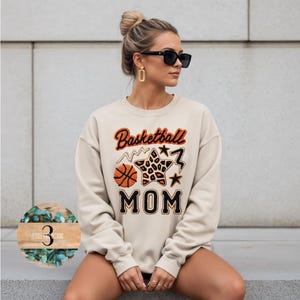 May include: A beige sweatshirt with the words "Basketball MOM" in orange and black lettering. The design includes a basketball, a star with a leopard print pattern, and star accents. The sweatshirt is a casual style.