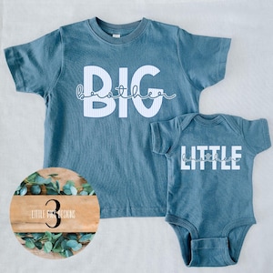May include: Two-piece set of matching blue shirts. One is a short-sleeved t-shirt with the words "BIG brother" in white. The other is a short-sleeved onesie with the words "LITTLE brother" in white. The shirts are made of a soft material.