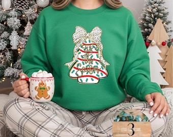 Pode incluir: Camisola verde com um desenho de sino de Natal que diz "Tis The Season". O sino tem um laço de bolinhas brancas e pretas. Uma caneca com um homem de gengibre é segurada na mão. A camisola é combinada com calças xadrez.