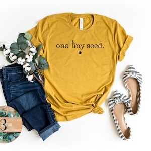 May include: A mustard yellow t-shirt with the text "one tiny seed." printed in black. The shirt is laying flat on a white surface with a pair of blue jeans, a sprig of eucalyptus, and a pair of striped flats.