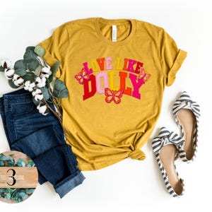 May include: A mustard yellow short-sleeve t-shirt featuring the text LOVE LIKE DOLLY in pink, red, and orange block letters, accented by three colorful butterflies. The shirt is paired with dark blue denim jeans and striped pointed-toe flats.