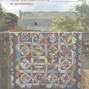 Könnte beinhalten: Ein farbenfroher Quilt mit einem Patchwork-Muster aus Kreisen und Quadraten. Der Quilt besteht aus verschiedenen Stoffen in Rot-, Blau-, Grün- und Gelbtönen. Der Quilt hängt an einem Holzzaun mit einer Backsteinmauer im Hintergrund. Der Titel "THE CIRCLE GAME" und der Text "BY JEN KINGWELL" sind oben im Bild gedruckt.