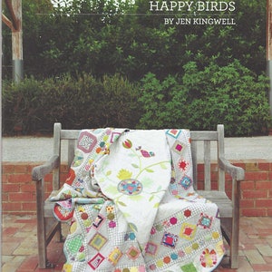 Könnte beinhalten: Eine farbenfrohe Patchwork-Decke mit weißem Hintergrund und einem Blumendesign mit Vögeln. Die Decke ist gefaltet und über eine Holzbank im Freien gelegt. Der Titel "Happy Birds" und der Name "Jen Kingwell" sind auf der Decke aufgedruckt.