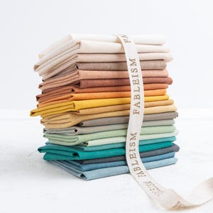 Può includere: Una pila di quadrati di tessuto piegati in un gradiente di colori, dal crema al turchese. Un nastro color crema con la scritta "FABLEISM" è avvolto attorno al tessuto. Il tessuto sembra essere cotone o lino.