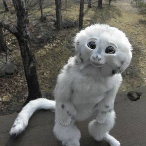 May include: A white and grey plush toy with large, black eyes and a long, fluffy tail. The toy is posed sitting up with its legs crossed and its head tilted to the side.