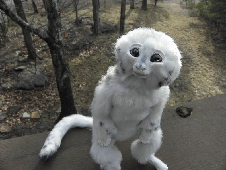 May include: A white and grey plush toy with large, black eyes and a long, fluffy tail. The toy is posed sitting up with its legs crossed and its head tilted to the side.
