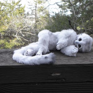 May include: A white plush toy with a long tail, lying on a wooden surface. The toy has large, black eyes and a fluffy, white body.
