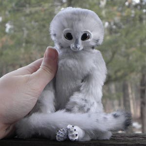 May include: A white plush toy with large, black eyes and a long, fluffy tail. The toy is sitting on a person's hand.