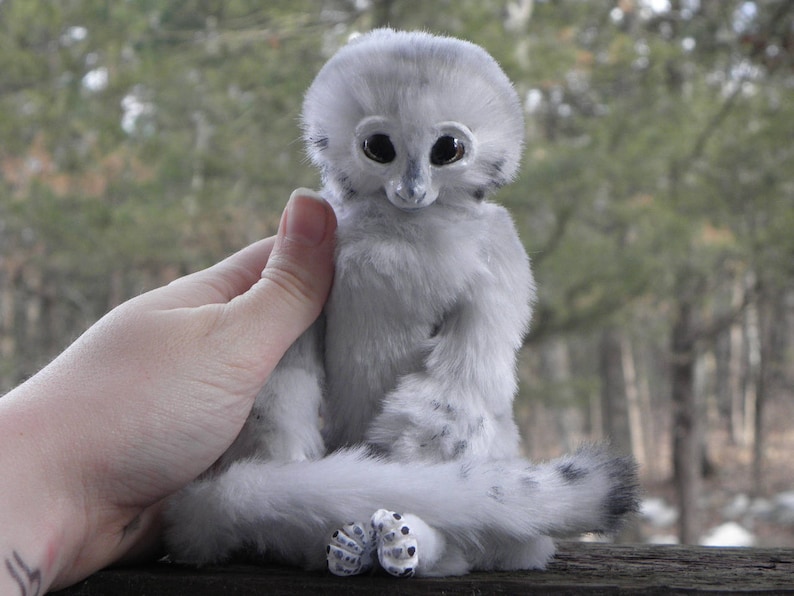 May include: A white plush toy with large, black eyes and a long, fluffy tail. The toy is sitting on a person's hand.