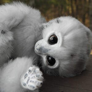 May include: A white, fluffy stuffed animal with large black eyes and a grey nose. The animal is lying on its side with its head turned to the side.