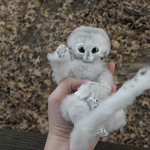 May include: A white plush toy resembling a lemur with large, black eyes and a long, bushy tail. The toy is being held by a hand.