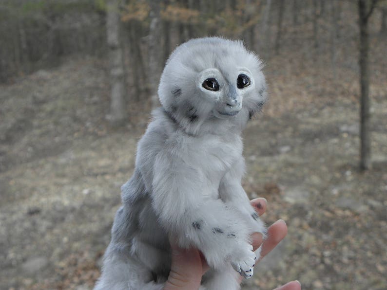 May include: A white, furry, stuffed toy with large, black eyes and a small, pink nose. The toy is sitting on a person's hand.