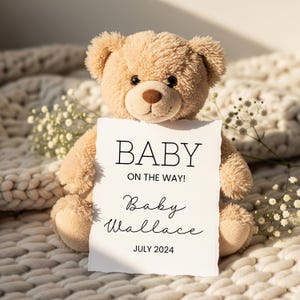 May include: A plush, tan teddy bear holds a white card with the text "BABY ON THE WAY!" and the name "Baby Wallace" with the date "JULY 2024". The bear sits on a cream-colored, textured blanket, with small white flowers in the background.