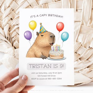 Puede incluir: Una invitación de cumpleaños con un capibara con un gorro de fiesta, globos y una tarta con velas encendidas. El texto dice "IT'S A CAPY BIRTHDAY!" y "TRISTAN IS 9!" con detalles del evento. La invitación es sostenida por una mano.