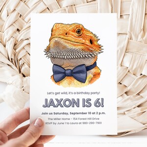 May include: Birthday party invitation featuring a cartoon illustration of a bearded dragon wearing a blue bow tie. The invitation reads "JAXON IS 6!" with event details. The invitation is held by a hand.