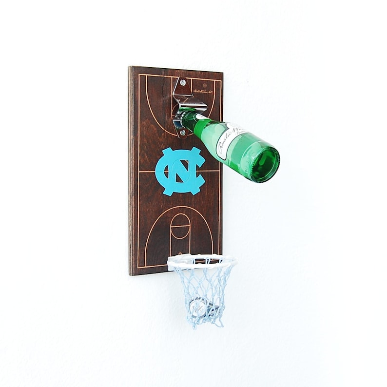 May include: A wooden basketball hoop with a bottle opener and a blue and white UNC logo. The hoop has a white net and a bottle is being opened in the opener.