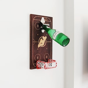 Puede incluir: Un abridor de botellas y una red de hockey montados en la pared. La madera marrón oscuro presenta un diseño de pista de hockey y un logotipo dorado. Se está abriendo una botella verde y debajo hay una red roja y blanca.