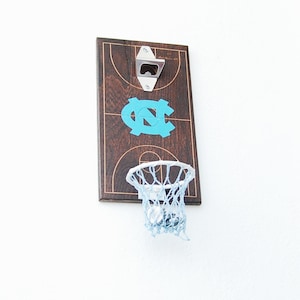 May include: A wooden basketball hoop bottle opener with a blue UNC logo on a brown basketball court background. The hoop has a white rim and a white net.