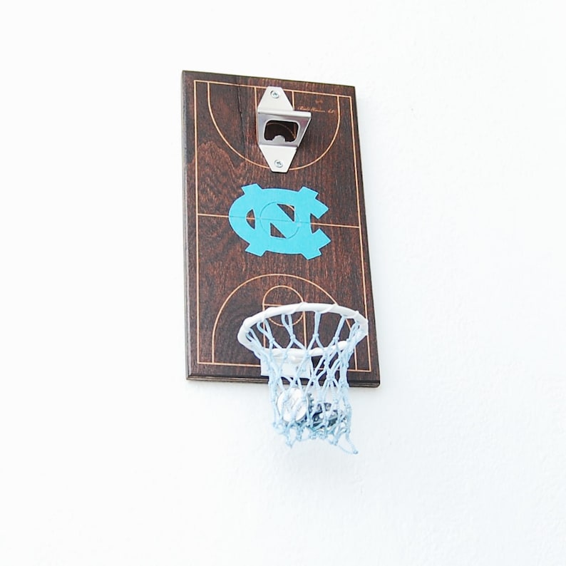 May include: A wooden basketball hoop bottle opener with a blue UNC logo on a brown basketball court background. The hoop has a white rim and a white net.