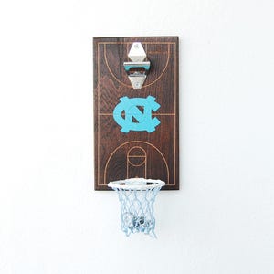 May include: A wooden bottle opener with a basketball court design and a mini basketball hoop. The court features the University of North Carolina Tar Heels logo in blue.