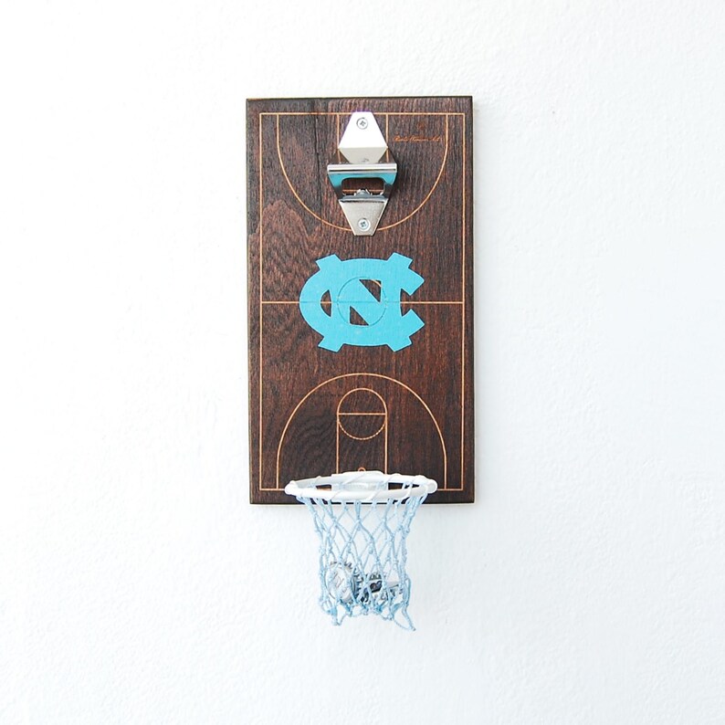 May include: A wooden bottle opener with a basketball court design and a mini basketball hoop. The court features the University of North Carolina Tar Heels logo in blue.