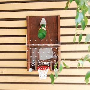 May include: A wall-mounted wooden game board featuring a dark brown finish, a metal bottle opener at the top, and a clear acrylic panel with the word Score printed in white. A small red and white basketball hoop is attached at the bottom center.