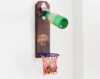 May include: A wall-mounted dark wood bottle opener featuring a silver metal opener mechanism at the top, a New York Knicks logo engraved in the center, and a small blue and orange basketball hoop with a net at the bottom catching bottle caps.