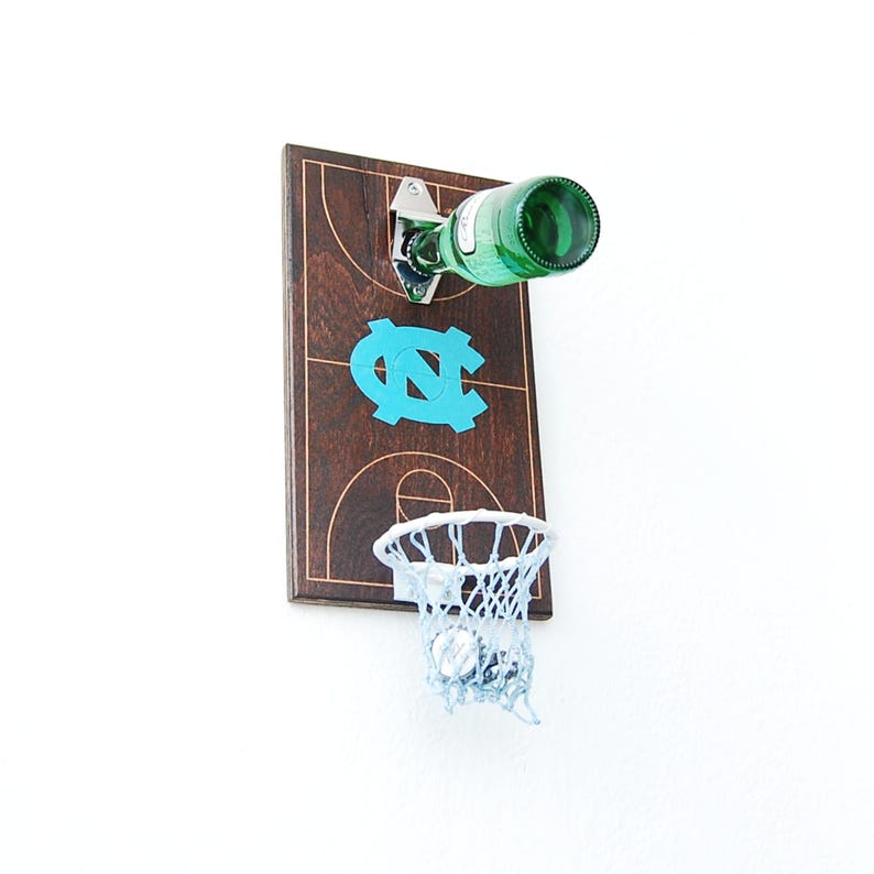 May include: A wooden basketball hoop with a bottle opener and a blue and white UNC logo. The hoop is mounted on a white wall and has a white net. A green glass bottle is hanging from the bottle opener.