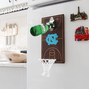 May include: A wooden bottle opener shaped like a basketball court with a net and a bottle opener attached. The court is brown with a blue and white logo in the centre. The bottle opener is attached to a white fridge.