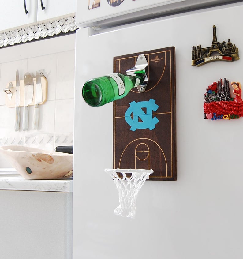 May include: A wooden bottle opener shaped like a basketball court with a net and a bottle opener attached. The court is brown with a blue and white logo in the centre. The bottle opener is attached to a white fridge.