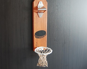 Op de afbeelding: Een houten wandbevestigde flessenopener met een basketbalring en een net eronder. De flessenopener heeft een zilveren metalen opener en een zwart silhouet van een flessenstop.