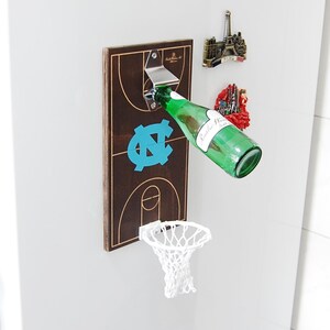May include: A wooden basketball hoop with a bottle opener and a white net. A green glass bottle is being opened with the bottle opener. The basketball hoop has a blue and white UNC logo.