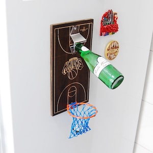 Può includere: Un apribottiglie in legno a forma di campo da basket, con il logo dei New York Knicks. Si sta aprendo una bottiglia verde. Un piccolo canestro da basket è attaccato sotto. I magneti decorativi sono visibili a destra.