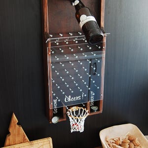 May include: A wooden wall-mounted bottle opener with a basketball hoop and a clear acrylic backboard. The backboard has a grid of screws and the text "Cheers!" printed on it. There are two bottle caps on the bottom left and right sides of the hoop. The bottle opener is holding a brown glass bottle.