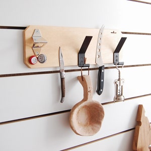 Op de afbeelding: Een houten wandorganizer met haken, een flesopener en een houten lepel. Er hangt een mes, een klein zakmes en een kurkentrekker aan de haken.