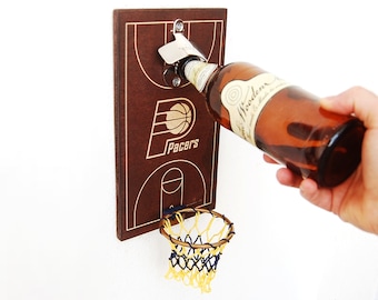 Puede incluir: Abrebotellas de madera en forma de aro de baloncesto con acabado de madera marrón y un abrelatas de metal. El aro tiene una red y el tablero tiene un diseño de cancha de baloncesto con las palabras "Pacers" impresas.