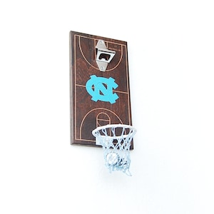 May include: A wooden bottle opener with a basketball hoop and net. The hoop is white and the net is white. The bottle opener is painted to look like a basketball court with the UNC Tar Heels logo in blue.