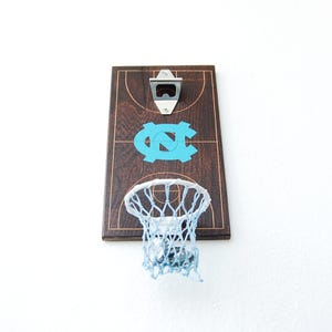 May include: A wooden bottle opener with a basketball hoop and net. The hoop is attached to a wooden board with a basketball court design and the letters "UNC" in blue. The bottle opener is silver.