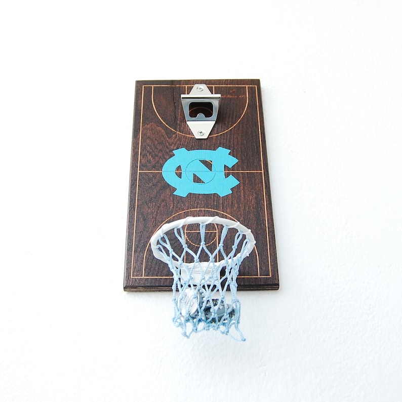 May include: A wooden bottle opener with a basketball hoop and net. The hoop is attached to a wooden board with a basketball court design and the letters "UNC" in blue. The bottle opener is silver.