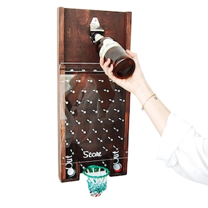 May include: A wall-mounted wooden bottle opener game featuring a clear acrylic front panel with a grid of metal pegs. The board includes the text Score, Out, and Out, with a small green and white basketball hoop attached at the bottom center.