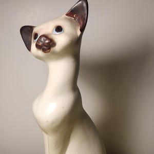 May include: A ceramic Siamese cat figurine in a seated position. The cat has a cream-coloured body, dark brown ears, paws, and nose, and blue eyes. The figurine is on a yellow surface.