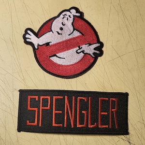 May include: Two embroidered patches. The top patch features the Ghostbusters logo: a white ghost inside a red circle with a red slash. The bottom patch is black with the name "SPENGLER" in red.