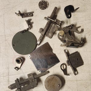 K&ouml;nnte beinhalten: Eine Ansammlung von Vintage-N&auml;hmaschinen-Teilen, darunter N&auml;hf&uuml;&szlig;e, Spulen und Platten, auf einer hellen Oberfl&auml;che. Die Metallkomponenten weisen Gebrauchsspuren auf und haben verschiedene Formen und Gr&ouml;&szlig;en.