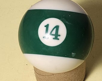 May include: A billiard ball, numbered 14, with a white top and bottom and a green stripe in the middle. The number 14 is in a white circle. The ball is resting on a small, cylindrical, brown support.