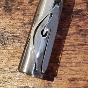May include: A silver metal bottle stopper with a pointed end and a decorative cut-out design. The stopper is cylindrical with a curved, flowing pattern cut into its side. It rests on a wooden surface.
