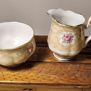 Op de afbeelding: Een suikerpot en roomkan set van crèmekleurig porselein met een gouden rand en een bloemenpatroon. De set heeft een delicaat bloemenpatroon met roze, rode en gele bloemen.