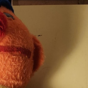 Op de afbeelding: Oranje pluche speelgoed met een blauwe hoed, een blauwe broek en een geel-groen gestreept shirt. Het speelgoed heeft een rode neus en een vriendelijke uitdrukking.