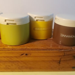 Könnte beinhalten: Vier vintage Gewürzgläser mit weißen Deckeln und Etiketten. Die Gläser sind orange, grün, gelb und braun. Die Etiketten lauten "NUTMEG", "GARLIC POWDER" und "CINNAMON". Die Gläser stehen auf einer Holzkiste.